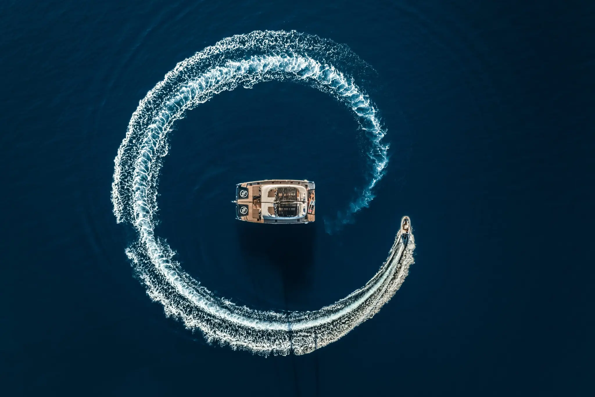 A crewed catamaran turning in deep blue water off a Greek island, seen from above