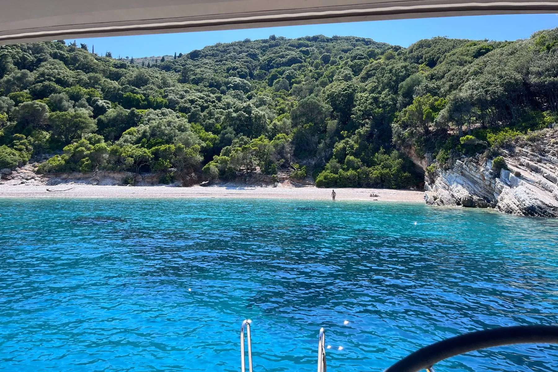 At anchor in a small bay - Oasis Sailing The view from a boat at anchor in a small Ionian bay, over the bow rail to a pebble beach and wooded shore