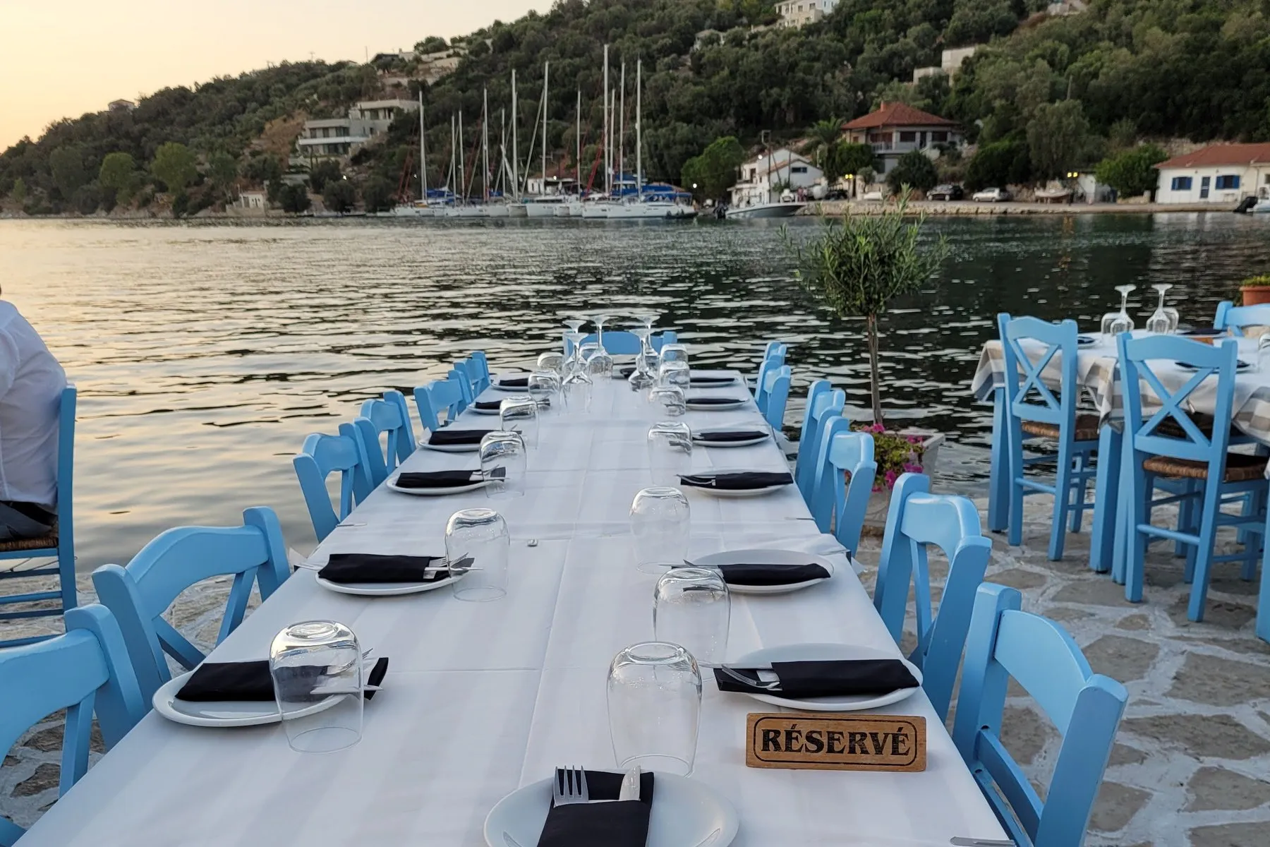 Dinner ashore - Oasis Sailing A long taverna table laid for a group beside the water, blue chairs down both sides and the masts of the quay behind