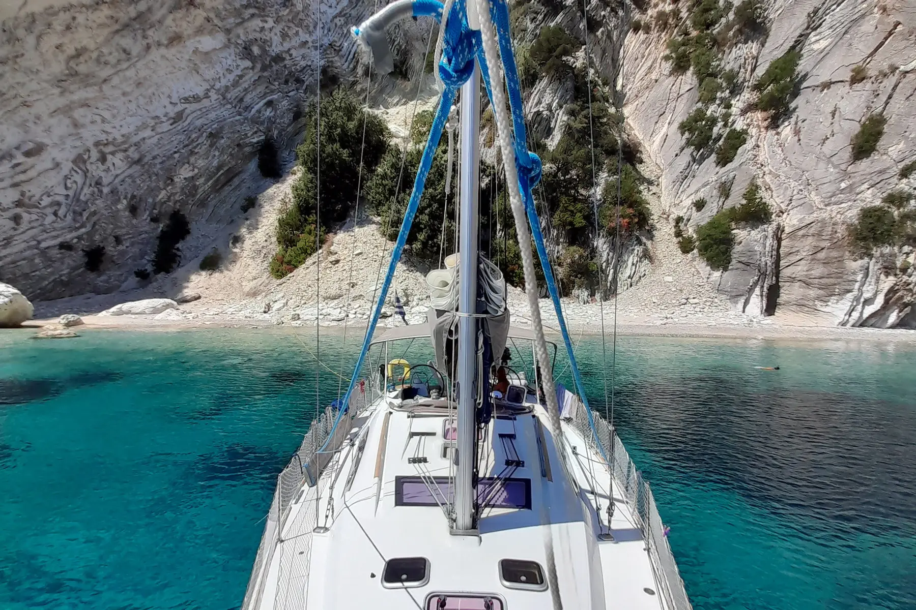 From the bow of a sailing yacht at anchor off a white-cliffed pebble beach, a swimmer in the turquoise water